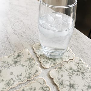 Könnte beinhalten: Ein Glas Wasser mit Eiswürfeln steht auf einem Untersetzer mit Blumenmuster. Drei weitere Untersetzer mit gewelltem Rand und cremefarbenem Hintergrund mit grauem Blumendesign sind sichtbar. Die Untersetzer haben einen Durchmesser von etwa 10 cm.