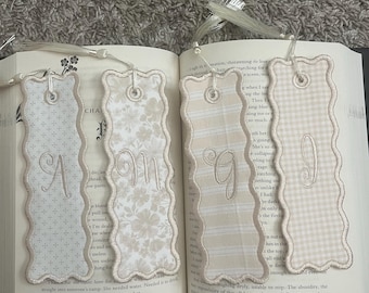 May include: Four rectangular fabric bookmarks with scalloped edges and white tassels. Each bookmark features a unique beige or cream pattern and a single embroidered letter: A, M, G, or I. The bookmarks are resting on an open book page.