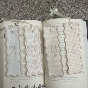 May include: Four rectangular fabric bookmarks with scalloped edges and white tassels. Each bookmark features a unique beige or cream pattern, including floral, cross, and gingham prints, with a single embroidered letter A, M, G, or I.