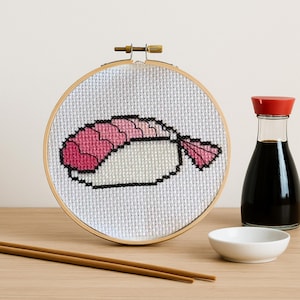 May include: A cross-stitch embroidery of a sushi nigiri in a wooden hoop. The sushi features pink and white details. A bottle of soy sauce, a small white bowl, and chopsticks are also in the image.