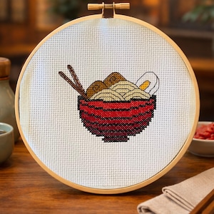May include: Cross-stitch embroidery of a bowl of ramen in a wooden hoop. The bowl is red with black stripes and contains noodles, egg, and meat. Chopsticks rest on top. The background includes a tea cup, a bottle, and a small bowl of red food.