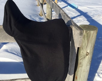 May include: Black saddle pad with a strap hanging from the bottom. The saddle pad is hanging on a wooden fence post in a snowy field.