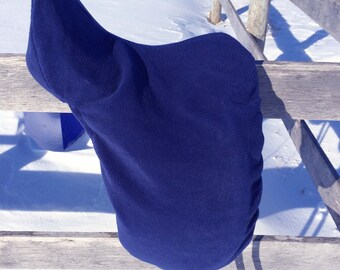 May include: A navy blue saddle cover is draped over a wooden fence in a snowy outdoor setting. The cover is made of a soft, textured material and has a rounded shape. Two black leather straps hang down from the cover.