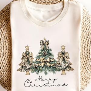 May include: Cream-colored t-shirt featuring three Christmas trees with leopard print bows and star toppers. The central tree has pinecones and a gold bow. The words "Merry Christmas" are written in script below the trees.