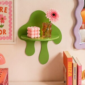 Op de afbeelding: Een groene paddenstoelvormige wandplank met een kleine ronde plank. De plank bevat een roze bubbelkubus, een kleine glazen vaas met een roze bloem en een ingelijste print met de tekst "MORE AMOR".