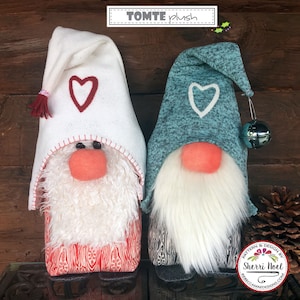 May include: Two plush Tomte gnomes, one with a white hat and red heart, the other with a teal hat and white heart. Both have white beards and are standing on a wooden surface. The text "TOMTE plush" is visible above the gnomes.