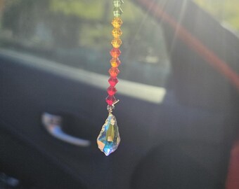 May include: A rainbow-colored crystal car charm. The charm is made of small, faceted beads that transition from red to blue, with a larger, teardrop-shaped crystal at the bottom. The charm hangs from a thin, clear string.