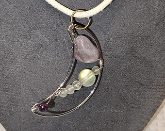 May include: A silver-toned crescent moon pendant necklace with a white cord. The pendant features a large, light purple stone, iridescent beads, and smaller purple and clear stones. The necklace is displayed against a gray background.