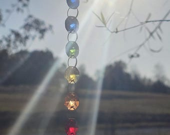 May include: A hanging suncatcher with a series of colorful, faceted crystal beads in a rainbow spectrum, suspended from a metal hook. The sun shines through the crystals, creating a sparkling effect. The bottom bead is a large, clear crystal.