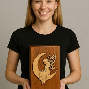 May include: A rectangular wooden box featuring a light-colored wood inlay of a fairy sitting on a crescent moon. The box is constructed from a medium-brown wood with a visible grain pattern, and the fairy figure is depicted in profile with wings.
