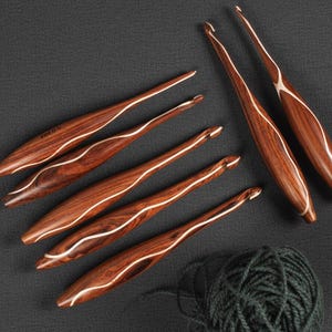 Peut inclure: Ensemble de crochets faits à la main en bois foncé avec une incrustation blanche. Ils sont disposés sur une surface gris foncé, avec une pelote de fil vert foncé en bas à droite. Les crochets ont une conception ergonomique incurvée.