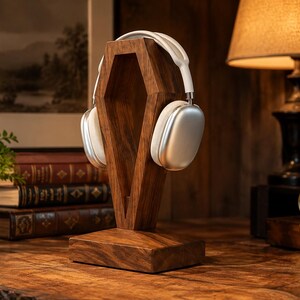 May include: A dark brown wooden headphone stand shaped like a coffin, featuring a rectangular base and a vertical support structure. A pair of silver over-ear headphones with a white headband is resting on the top of the wooden stand.
