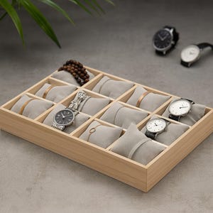 May include: A wooden watch and jewelry organizer with multiple compartments. The organizer contains various watches, bracelets, and rings displayed on gray velvet pillows. The watches have black and white faces and leather or metal bands.