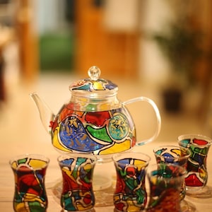 May include: A clear glass teapot and six matching glasses, all adorned with vibrant, stained-glass-style designs in red, blue, yellow, and green. The teapot features a handle and a lid with a decorative knob. The glasses are small and elegant.
