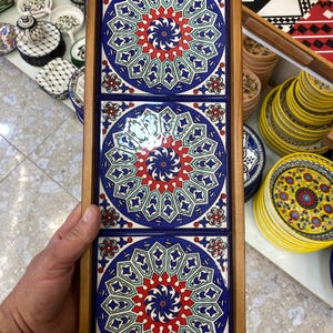 May include: A wooden tray holding three square ceramic tiles. Each tile features a circular design with a blue background, red and white accents, and a central spiral pattern. The tray is made of light-colored wood.