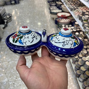 Peut inclure: Un plat à condiments en céramique à deux compartiments avec couvercles, tenu à la main. Le plat est blanc avec des bordures bleues et présente des motifs floraux et d'oiseaux colorés. Les couvercles ont un petit bouton sur le dessus.