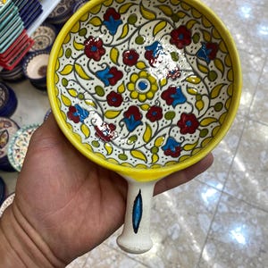 Può includere: Un piatto da portata in ceramica dipinto a mano con bordo e manico gialli. Il piatto presenta un interno bianco con un motivo floreale rosso, blu e giallo. Il manico è bianco con un accento blu.