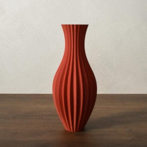 Puede incluir: Un jarrón de color terracota con un diseño vertical acanalado. El jarrón tiene un cuello delgado y una base ancha, sobre una superficie de madera. El jarrón mide aproximadamente 25 cm de alto.