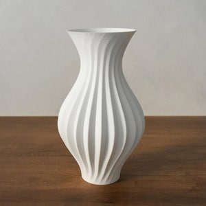 Puede incluir: Un jarrón blanco con un diseño texturizado y acanalado. El jarrón tiene una base ancha que se estrecha hacia un cuello estrecho y una abertura acampanada. El jarrón está sobre una superficie de madera.