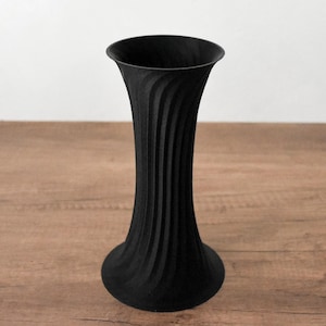 Può includere: Un vaso nero alto con la parte superiore svasata e una base arrotondata. Il vaso presenta un design a spirale verticale strutturato. È realizzato in materiale nero opaco e poggia su una superficie di legno.