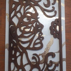 Può includere: Un'opera d'arte in legno con una silhouette di profilo intagliata in legno marrone scuro. Il profilo è posto su uno sfondo bianco con accenti di foglia d'oro. Il pezzo è rettangolare con un elemento floreale decorativo.