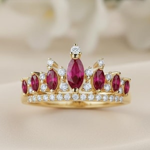 May include: A gold crown ring with ruby gemstones and small diamonds. The ring has a double band design. The rubies are marquise-cut. The ring is set against a soft, neutral background, highlighting its elegant details.