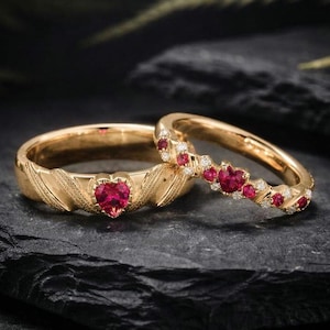 Peut inclure: Deux bagues en or. L'une présente un rubis en forme de cœur et des gravures en forme de feuilles. L'autre bague est ornée de rubis en forme de cœur et de petits diamants. Les deux bagues sont posées sur une surface sombre et texturée.