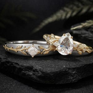 May include: Two rings with intricate designs. One features a pear-shaped diamond, the other a square-cut diamond. Both rings have gold leaf accents and are set on a dark surface, creating a striking contrast. The rings are likely engagement or wedding rings.