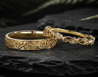 May include: Two gold wedding rings. The larger ring features an intricate floral and vine pattern. The smaller ring has a delicate design with small diamonds. Both rings are displayed on a dark, textured surface.