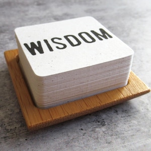 May include: A stack of square white cards with the word WISDOM printed in black capital letters, resting inside a square wooden holder. The cards have a speckled texture and rounded corners, and the holder features a natural wood grain finish.