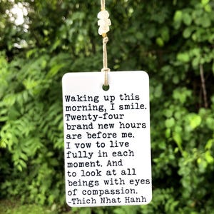 May include: A white rectangular tag with a quote by Thich Nhat Hanh, suspended by a beige string with small white beads. The text reads: "Waking up this morning, I smile. Twenty-four brand new hours are before me..."
