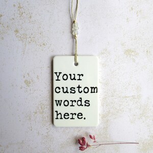 May include: A white rectangular tag with the text "Your custom words here" in black. The tag is suspended by a beige string with small beads. A sprig of dried red flowers rests below the tag.