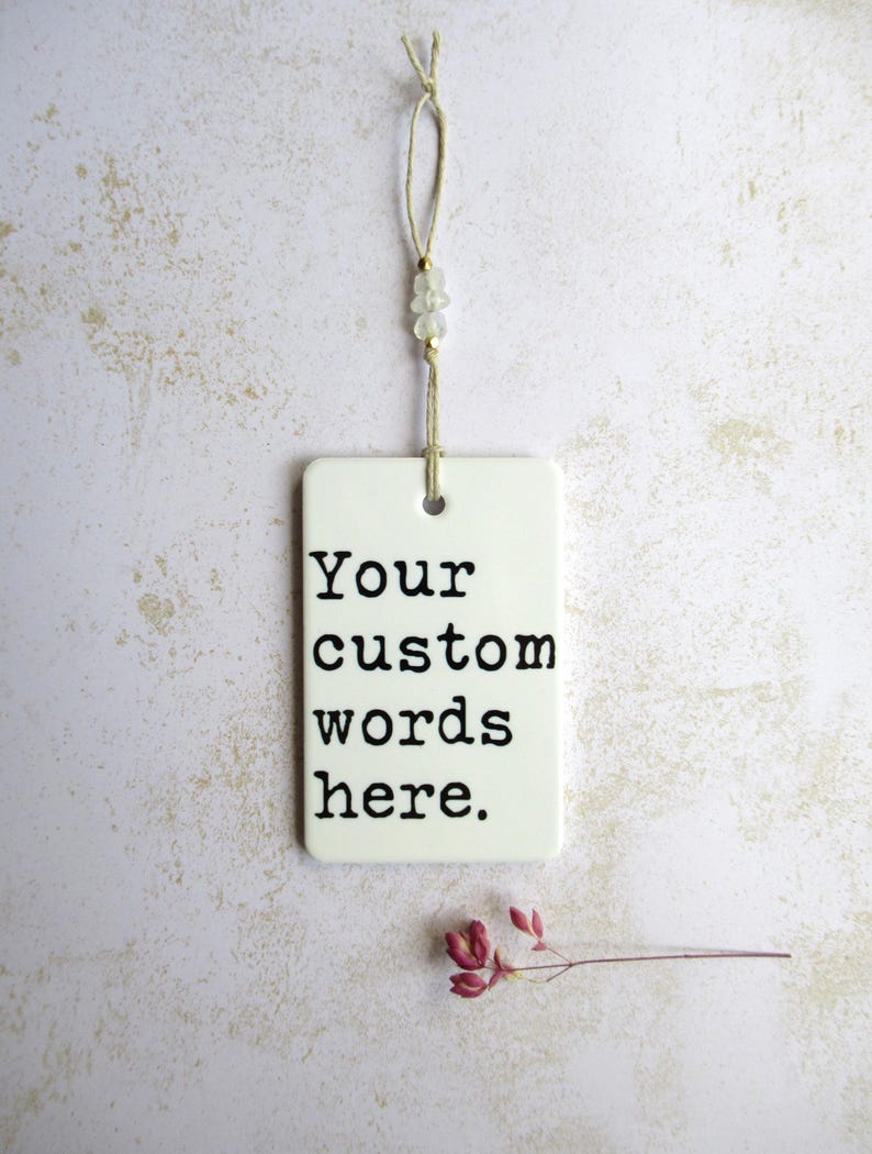 May include: A white rectangular tag with the text "Your custom words here" in black. The tag is suspended by a beige string with small beads. A sprig of dried red flowers rests below the tag.