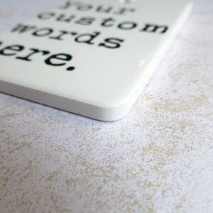 May include: A white rectangular tag with rounded corners, featuring the text "Your custom words here" in black. The tag is hanging by a thin, off-white string. The background is a textured, off-white surface.