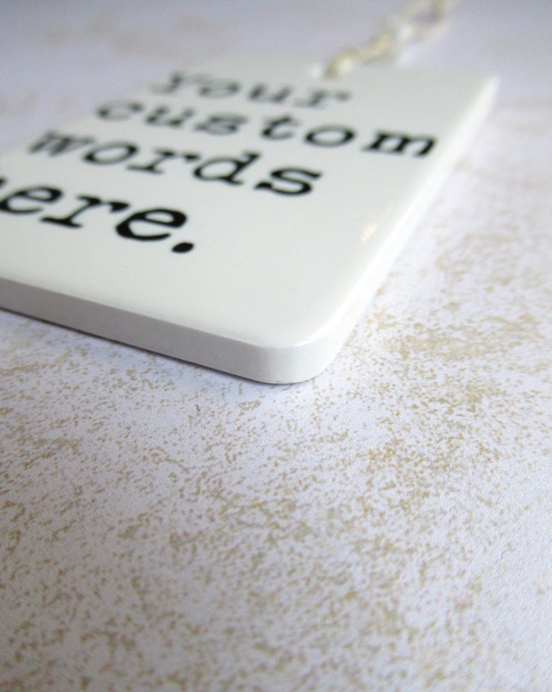 May include: A white rectangular tag with rounded corners, featuring the text "Your custom words here" in black. The tag is hanging by a thin, off-white string. The background is a textured, off-white surface.