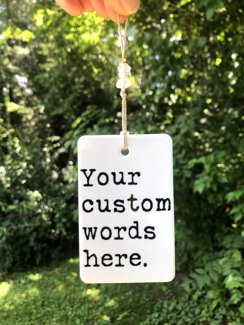 May include: A white rectangular tag with the text "Your custom words here" in black. The tag is suspended by a natural-colored cord adorned with small, clear beads. The background is a blurred view of green foliage.