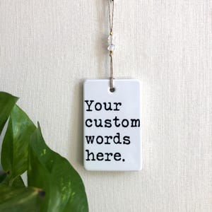 May include: A white rectangular ceramic tag with the text "Your custom words here" in black, hanging from a string with decorative beads. The tag is suspended against a textured white background.