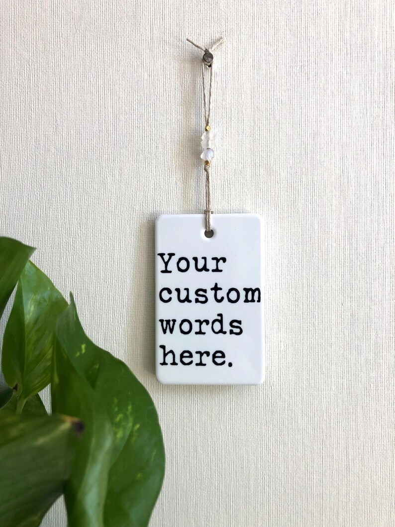 May include: A white rectangular ceramic tag with the text "Your custom words here" in black, hanging from a string with decorative beads. The tag is suspended against a textured white background.