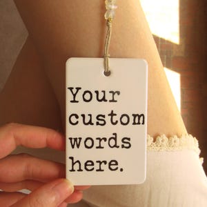 May include: A white rectangular tag with the text "Your custom words here" in black. The tag is suspended by a beige string with small, clear beads. The tag is held by a hand.