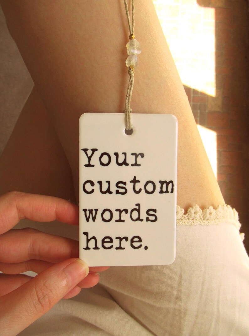May include: A white rectangular tag with the text "Your custom words here" in black. The tag is suspended by a beige string with small, clear beads. The tag is held by a hand.