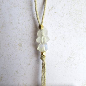 May include: A handmade necklace featuring three clear, rough-cut gemstones strung on a beige cord. The gemstones are separated by small, gold-colored beads. The cord is knotted at the top and bottom, creating a simple, elegant design.