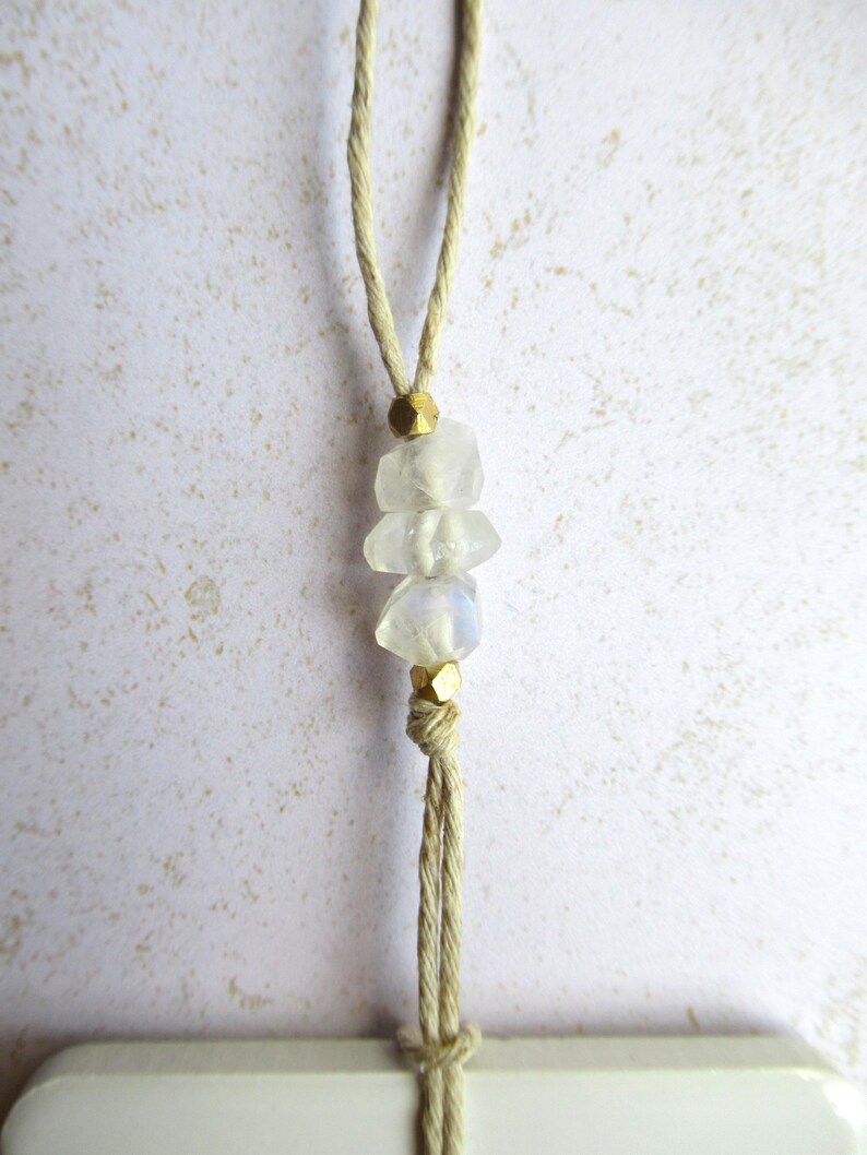 May include: A handmade necklace featuring three clear, rough-cut gemstones strung on a beige cord. The gemstones are separated by small, gold-colored beads. The cord is knotted at the top and bottom, creating a simple, elegant design.