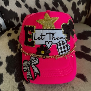 May include: A vibrant pink trucker hat adorned with gold glitter star, black bow, and checkered heart patches. The hat features the text "Let Them" and a gold chain. A stylish accessory.