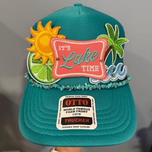 May include: A teal trucker hat featuring a central pink patch with the text IT'S Lake TIME, surrounded by embroidered patches of a yellow sun, a green lime slice, a palm tree, and blue waves, with a row of turquoise stones and an OTTO brand sticker.