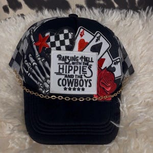 May include: Black trucker hat with a checkered pattern and a gold chain. The hat features patches with a skeleton hand, playing cards, a red star, a red rose, and the text "Raising Hell with the Hippies and the Cowboys."