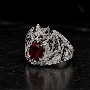 May include: A silver ring featuring a bat design with a large, oval, red gemstone at the center. The bat has black and white wings, and the ring is encrusted with small, clear gemstones.
