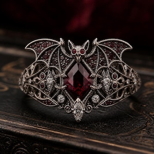 May include: A silver-toned ring featuring a bat design with ruby-red and clear gemstones. The ring has a large, square-cut ruby at its center, with smaller rubies and clear stones accenting the bat wings and band. The ring is set against a dark background.