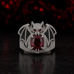 Front view of gothic vampire bat ring with oversized oval ruby red gemstone surrounded by black stone bat wings and silver pave detailing. Fantasy gothic statement ring inspired by vampire jewelry, dragon wing ring, Halloween fashion accessory, dark fantasy jewelry, gothic engagement ring, luxury cocktail ring, alternative style jewelry for men and women, spooky gothic gift, handmade silver gothic ring with dramatic vampire aesthetic.