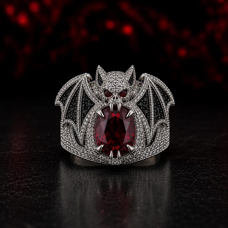 Front view of gothic vampire bat ring with oversized oval ruby red gemstone surrounded by black stone bat wings and silver pave detailing. Fantasy gothic statement ring inspired by vampire jewelry, dragon wing ring, Halloween fashion accessory, dark fantasy jewelry, gothic engagement ring, luxury cocktail ring, alternative style jewelry for men and women, spooky gothic gift, handmade silver gothic ring with dramatic vampire aesthetic.