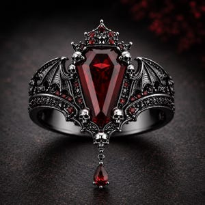 May include: A gothic-style ring with a large, rectangular, deep red gemstone at its center. The ring features intricate details, including a crown, skulls, and bat wings, all in a dark, metallic finish. A teardrop-shaped red gem hangs below.